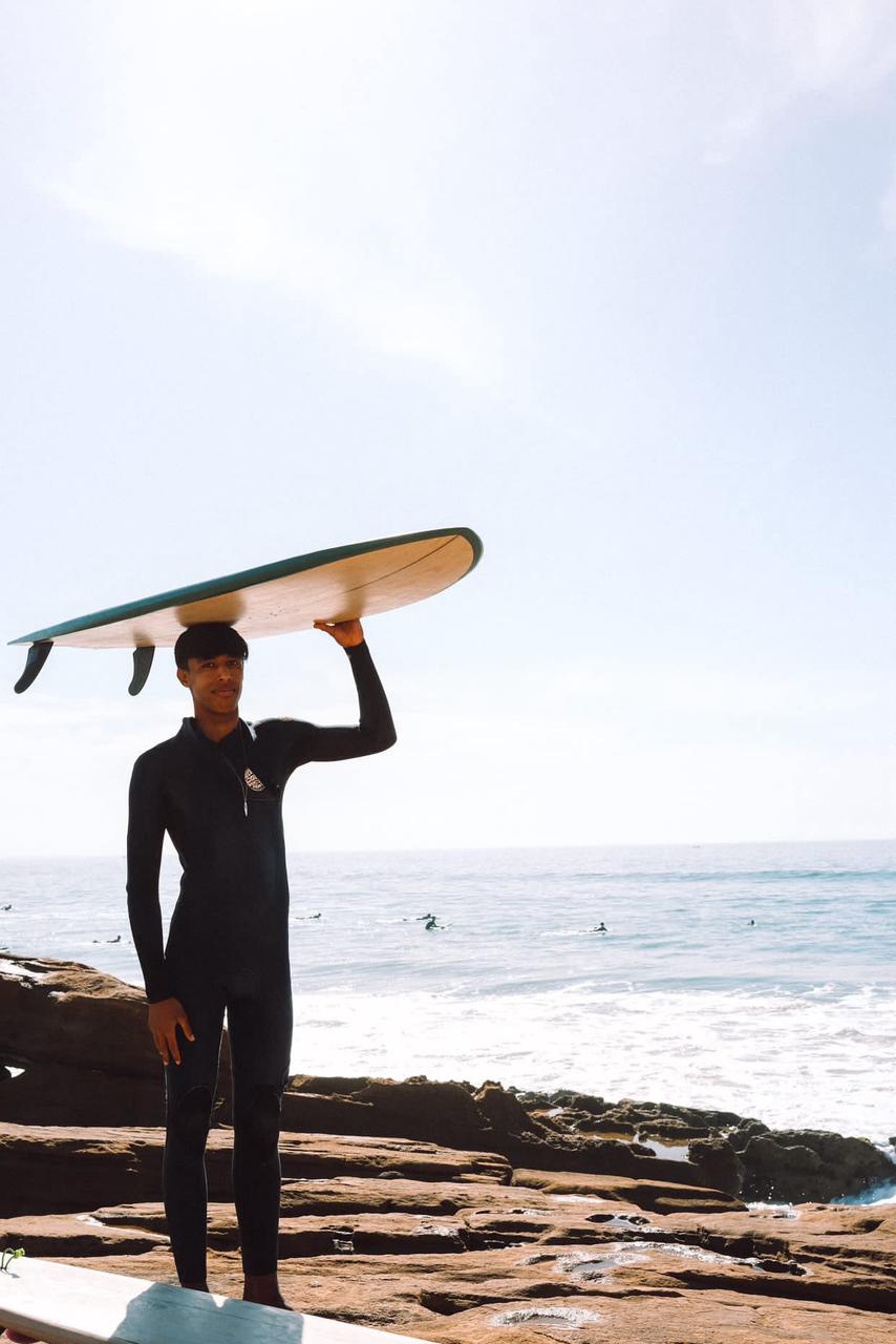Mohamed carrying his surfboard on his head on the rocks of Taghazout